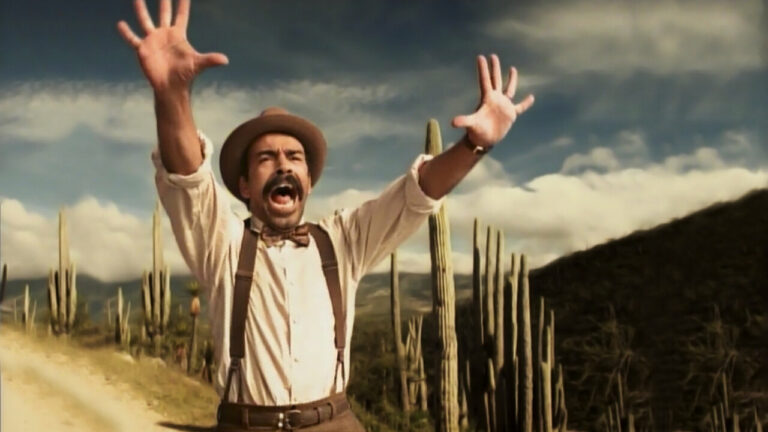 A man stands on a road lined with cacti. He stands with his hands up, yelling.