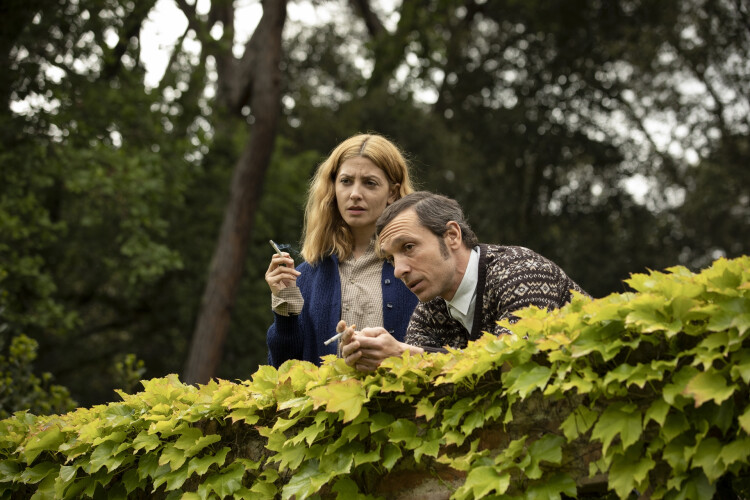 Film still of a man and a woman standing behind an ivy covered wall smoking cigarettes. Best Spanish movies to watch to learn the language.