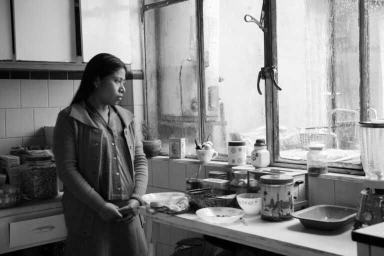 An image of a woman standing in a kitchen, looking sadly out the window.