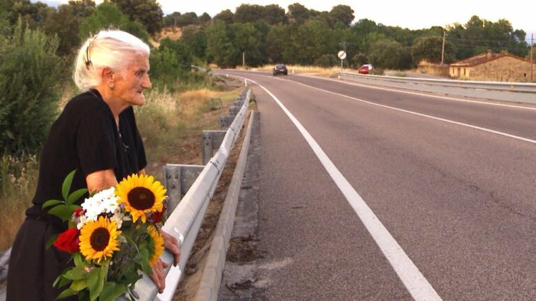 A white-haired woman stands next to a highway with a bouquet of flowers.