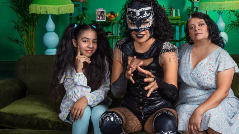 Image of two women and a girl sitting on a couch. One woman is in full lucha libre costume.
