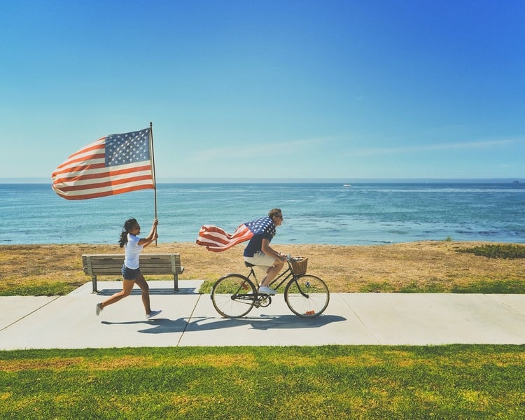 Seaside backdrop. Man biking wearing American flag like a cape, girl running after bicycle waving another American flag.
