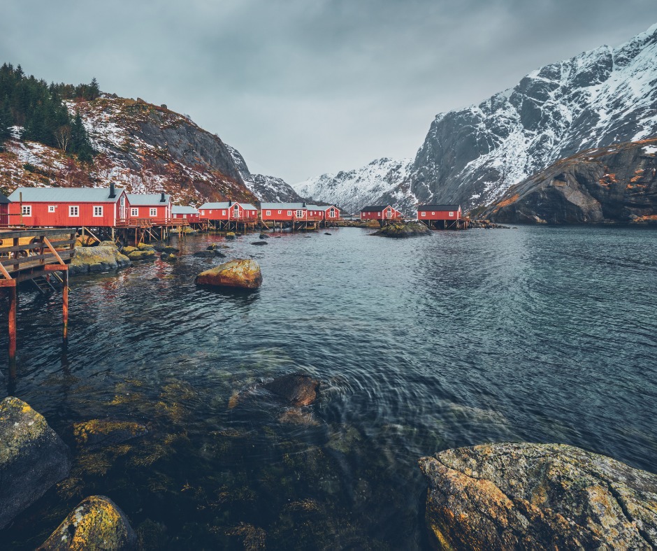 Red buildings overlooking the water and snowy mountains in Norway. Is Norwegian easy to learn for English speakers? It ranks 4th on our list!