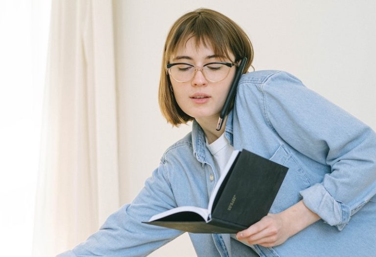 Women wearing glasses and a blue button down shirt, holding a phone against her ear with her shoulder and a planner in one hand.
