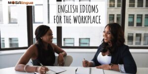 Two smiling professional Black women sitting at a table in front of a window with notebooks in front of them. Text reads: English idioms for the workplace. MosaLingua