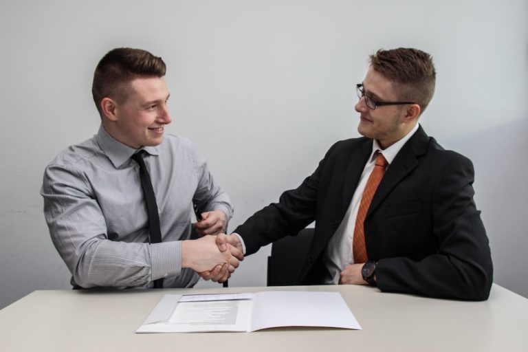 Two white men in business cuits sitting at a table in front of an open folder, smiling and shaking hands.