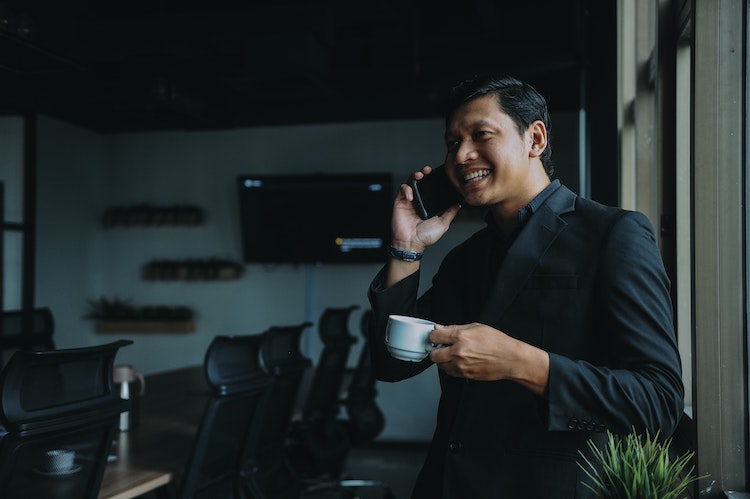 Man wearing an all-black business suit, smiling with a phone up to his ear and a coffee cup in the other hand, standing in front of a dark conference room.