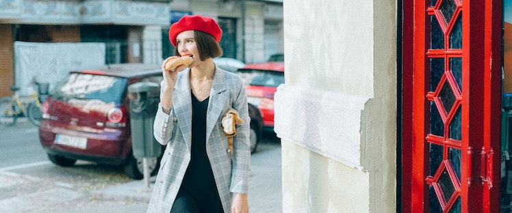 A chic woman with short brown hair, wearing a red beret and gray blazer and eating a baguette on the street in France.
