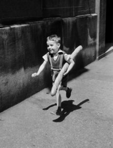 A little boy in a black and white photograph running down the street with a baguette.